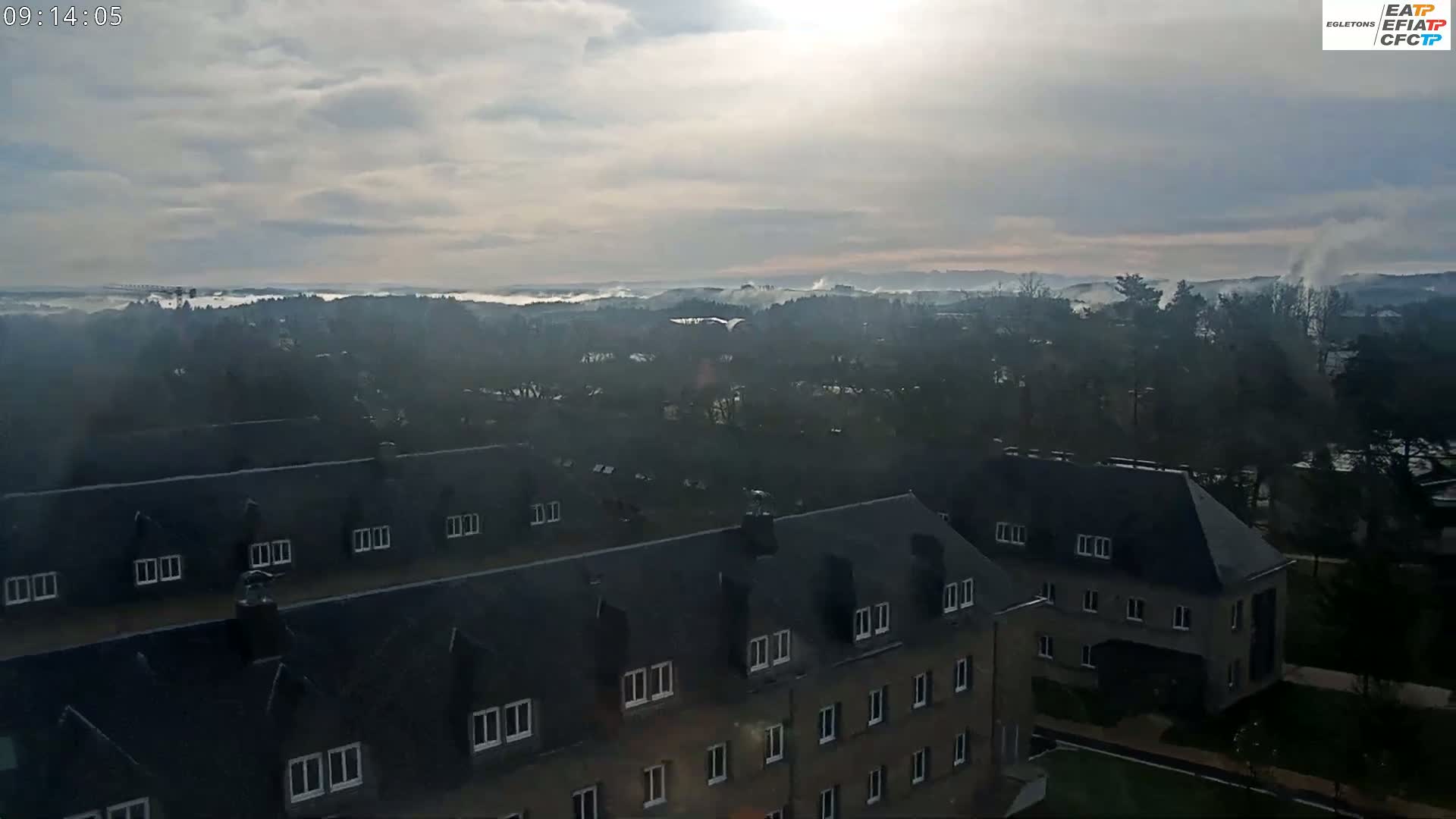 A hazy, overcast day reveals a low-lying fog over a tree-covered landscape, viewed from above a row of dark-roofed buildings.