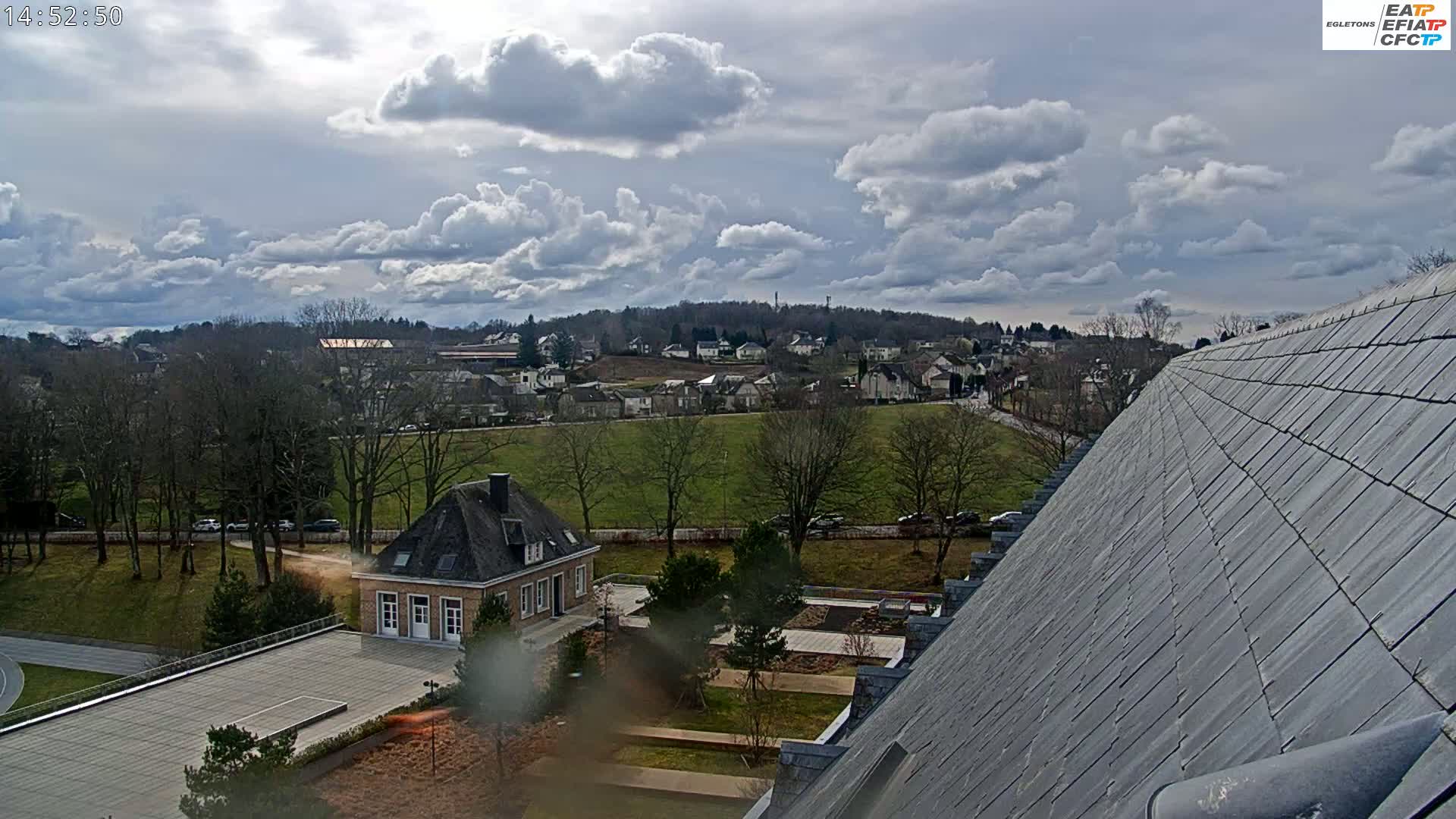 A partly cloudy sky overlooks a small town nestled among trees and a large, sloped roof, with a small building and courtyard visible in the foreground.