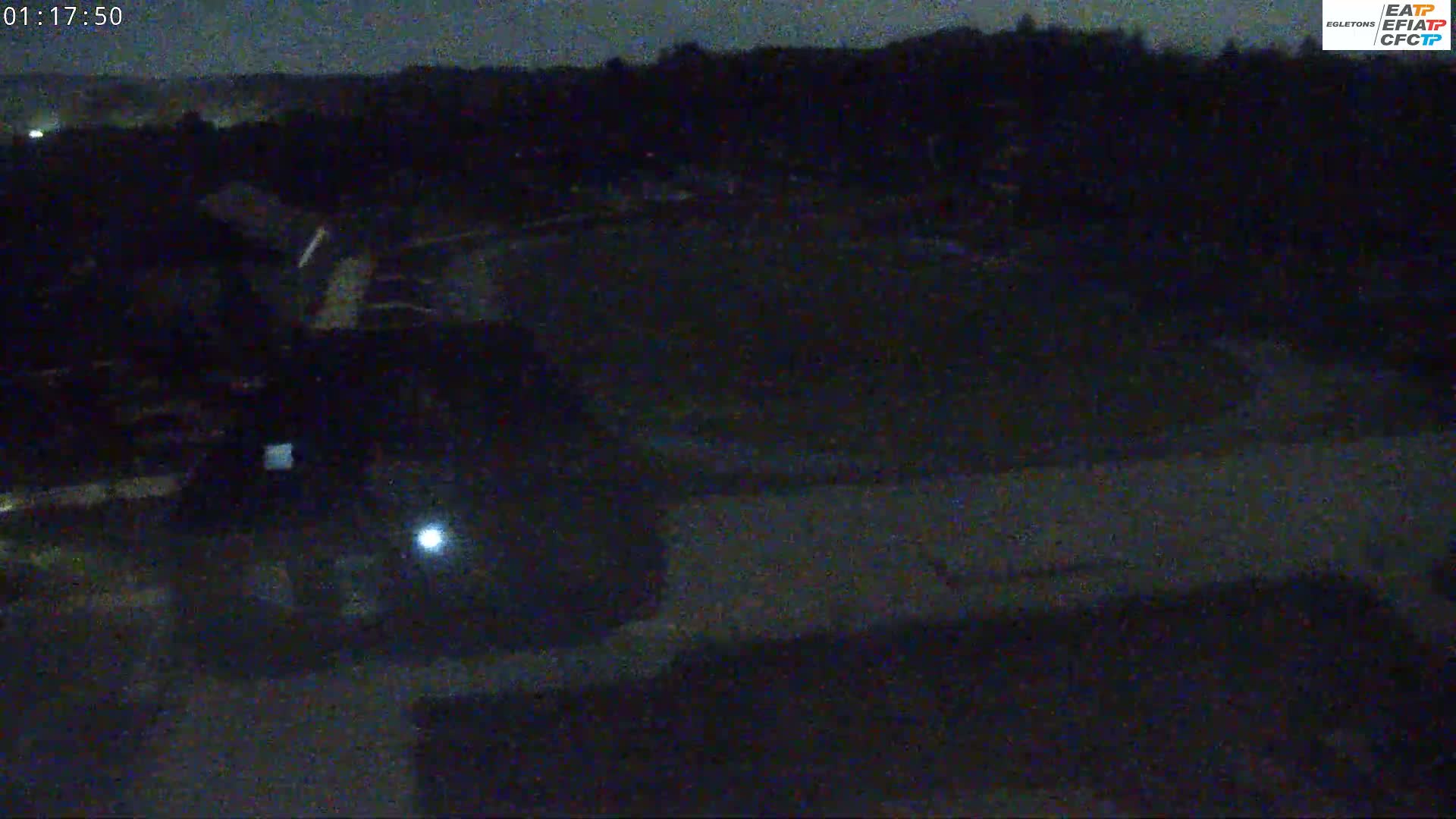 A nighttime, low-light view shows a dark, possibly paved area, surrounded by dark buildings and trees under a dark sky.