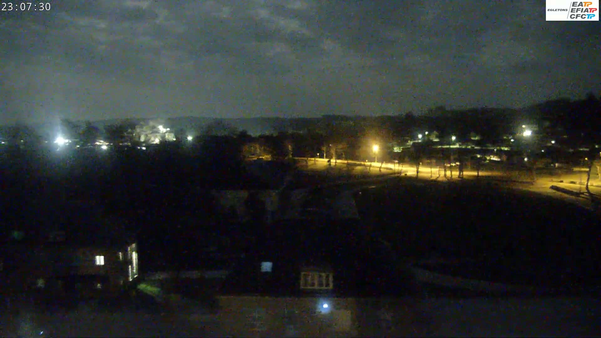 A nighttime view of a mostly dark landscape with some distant lights and a few illuminated buildings under a cloudy sky.