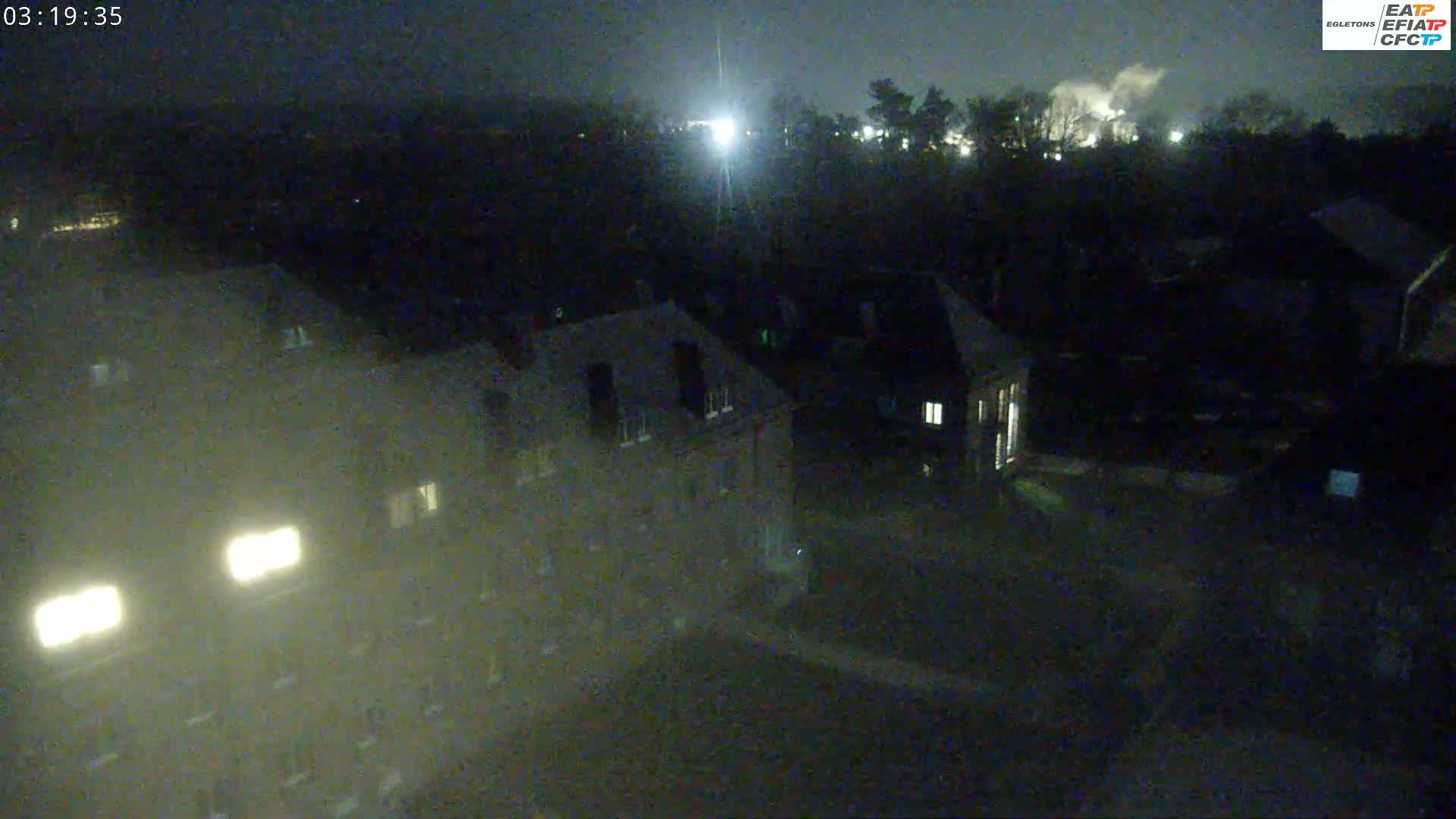 A nighttime, high-angle view shows several buildings with dimly lit windows, surrounded by dark trees and foliage under a dark sky.