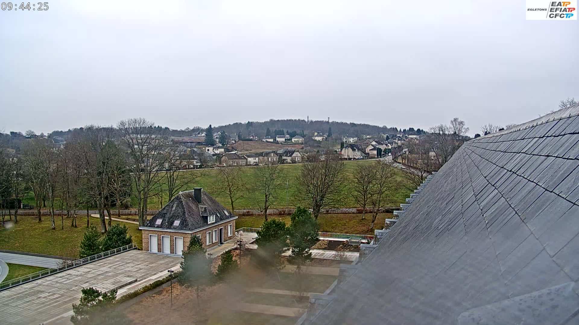 A view from a high vantage point shows a small town nestled in a valley under an overcast sky, with a grey slate roof dominating the foreground and a small building with a large paved area visible in the midground.