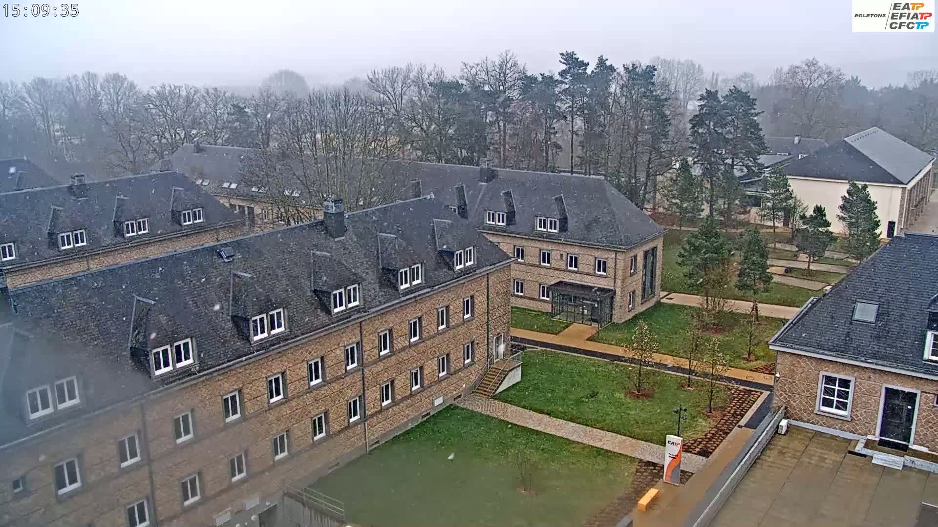 An aerial view shows several low brick buildings with dark roofs under a light snowfall.