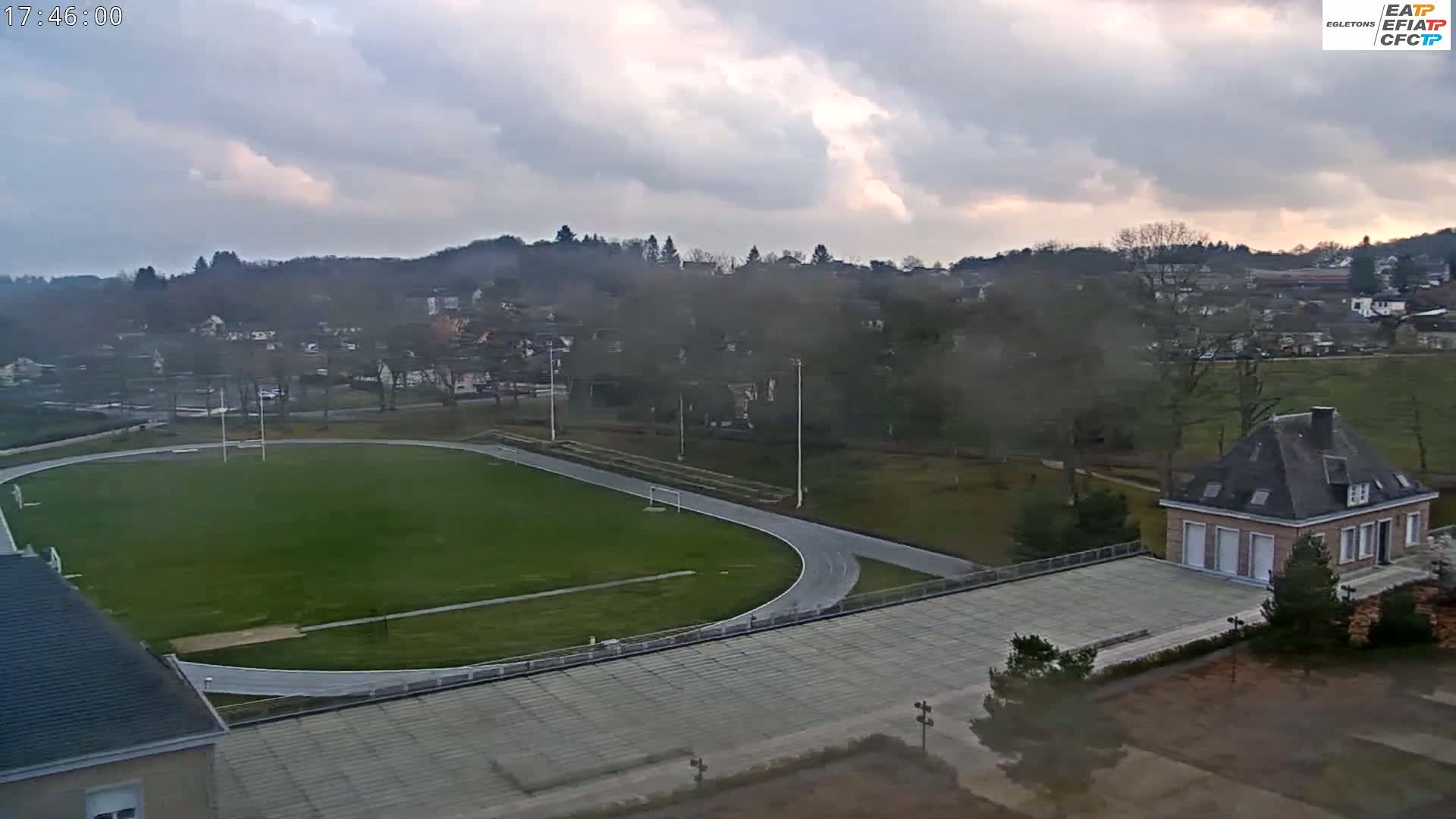An overcast day reveals a high-angle view of an oval running track encompassing a grassy field, situated near a small town nestled among hills, with a building to the lower right.