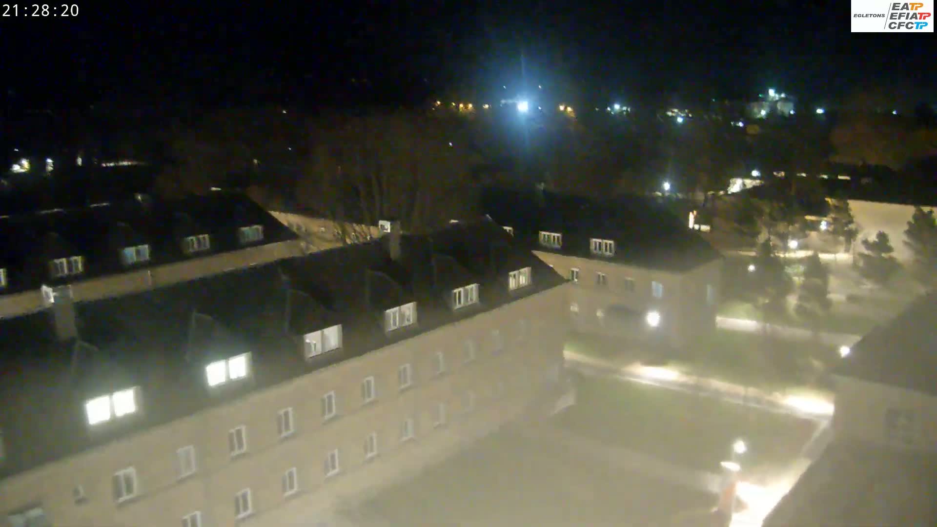 A nighttime aerial view shows a complex of low-rise buildings with illuminated windows, surrounded by sparsely lit grounds and trees.