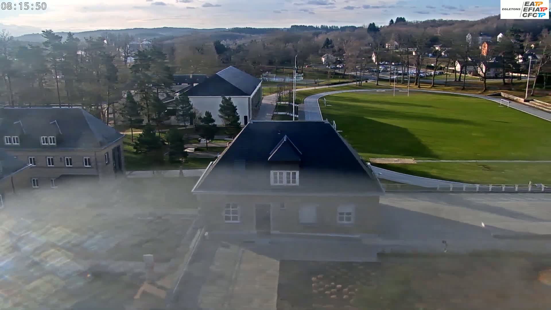 A high-angle, long shot shows a partly cloudy day overlooking a grassy field, track, and several buildings on a campus-like setting.