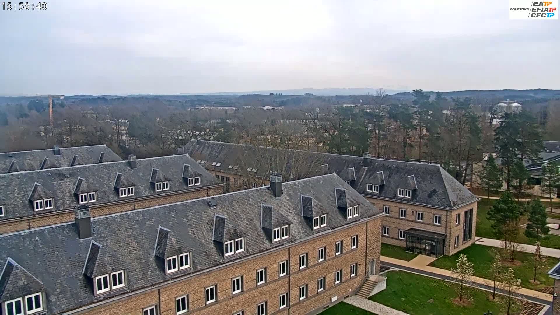 A large, long, low brick building with a gray slate roof is situated on a grassy campus overlooking a wooded area under an overcast sky.