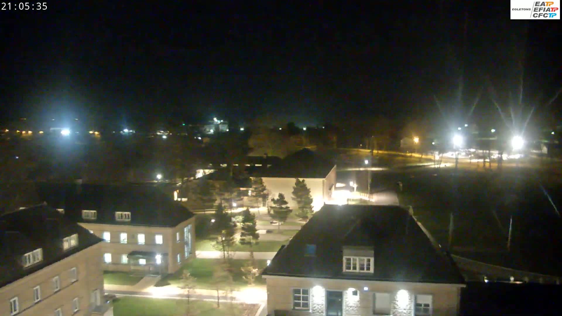 A nighttime, aerial view shows several illuminated buildings in a campus-like setting, surrounded by trees and paved areas.
