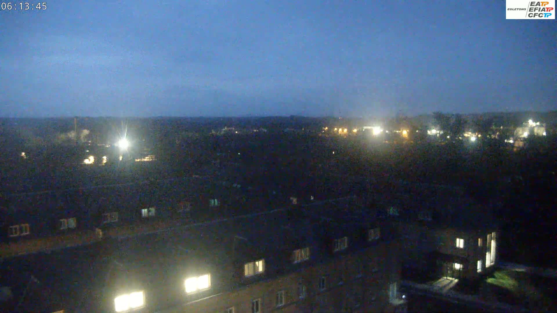 A nighttime, high-angle view shows a sprawling area of dark rooftops with a few lit windows, overlooking a dark landscape speckled with distant, faint lights under a twilight sky.