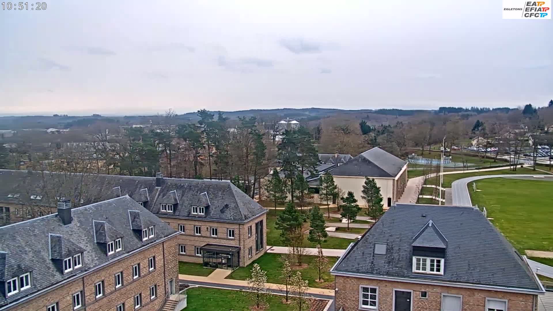 An aerial view of a campus with several low stone buildings under an overcast sky shows a running track and a grassy field in the background.