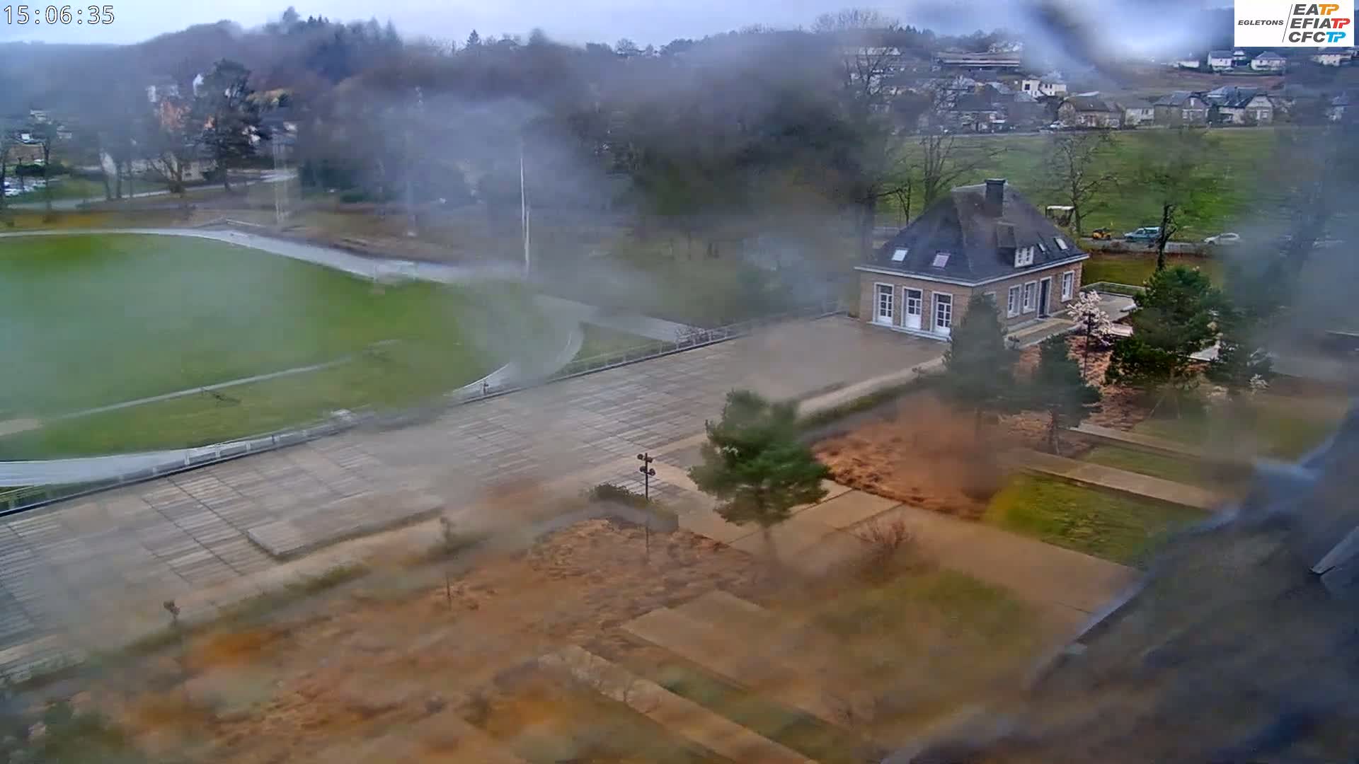 A high-angle view shows a rain-covered scene of a paved courtyard, a grassy athletic field, and a small stone building, all surrounded by trees and landscaping.