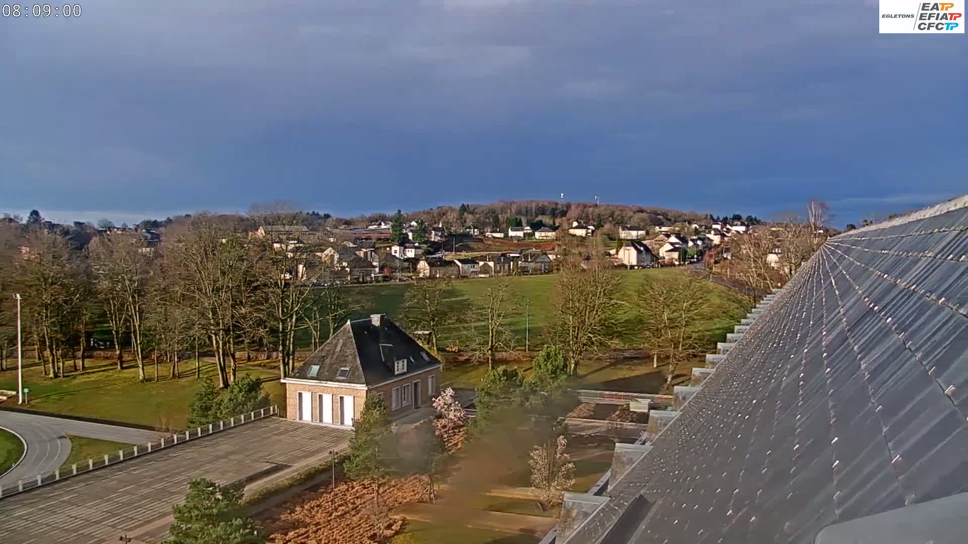 A small town nestled in a valley under a mostly clear blue sky is viewed from atop a sloped, dark gray roof.