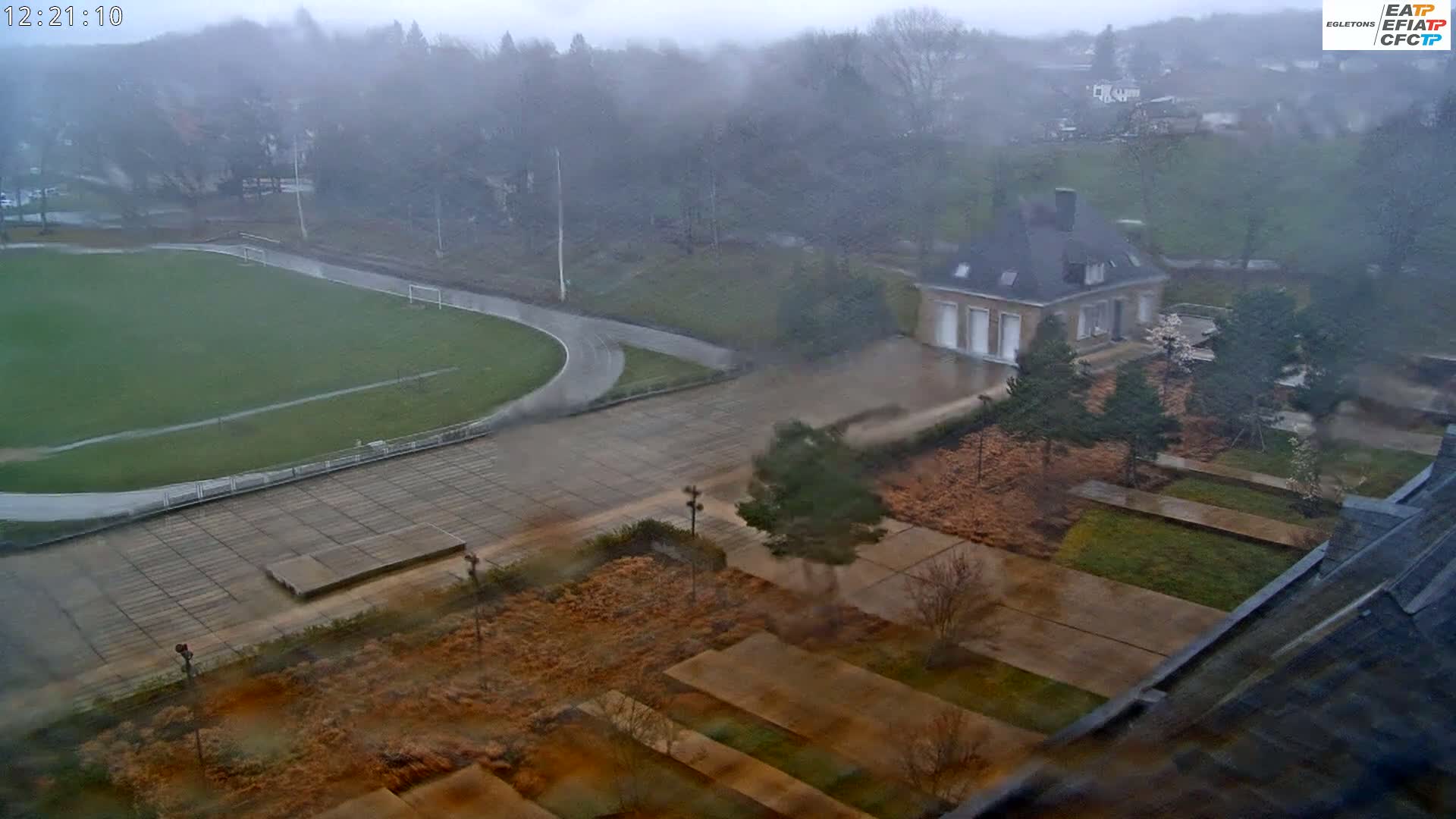 A high-angle view shows a rain-soaked athletic field, paved areas, and a building amidst landscaping, all under a heavy, overcast sky.