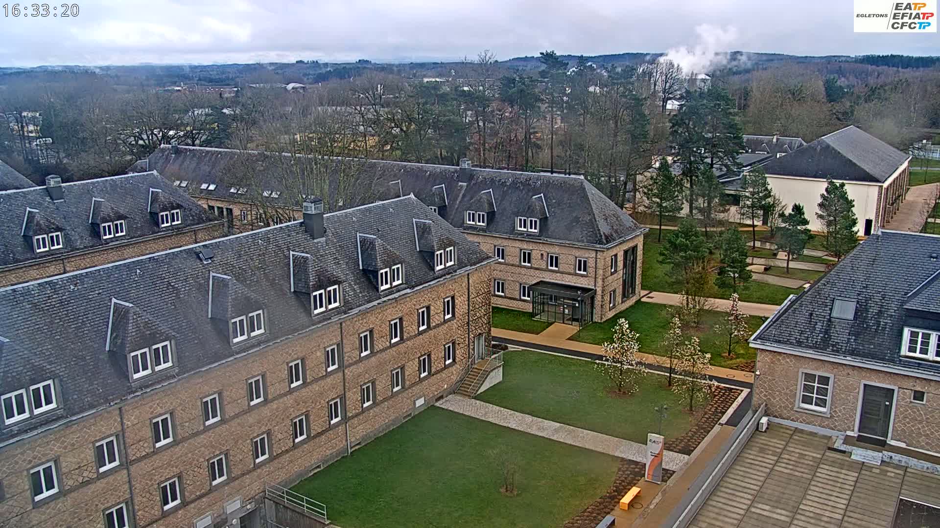 An aerial view shows several low, long, stone buildings with dark gray roofs on a partly cloudy day, surrounded by lawns, trees, and walkways.