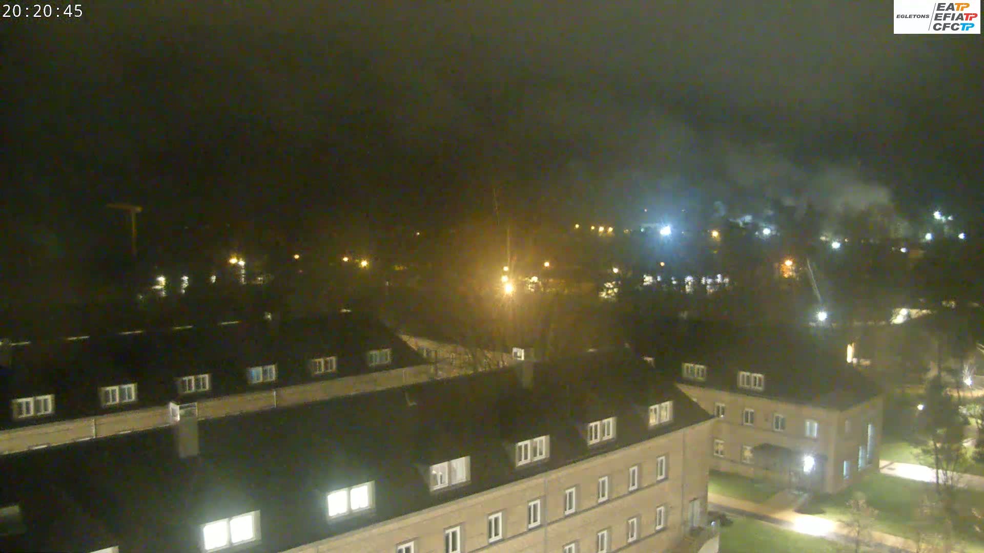 A nighttime, hazy view shows a complex of low-rise buildings with illuminated windows, surrounded by sparsely lit grounds and distant lights.