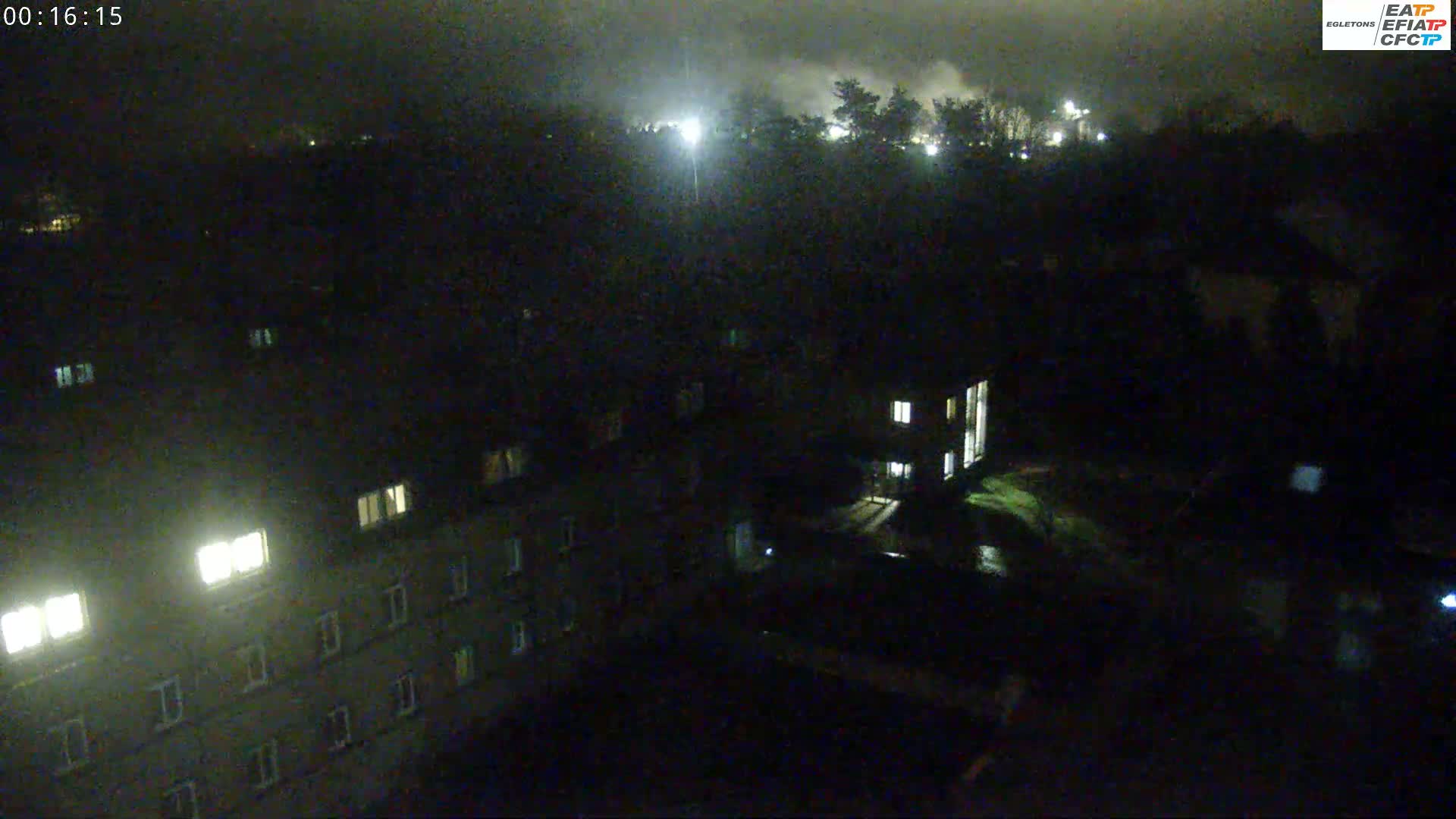 A nighttime aerial view shows a building complex with some lit windows, trees, and a distant light source, under a dark and cloudy sky.