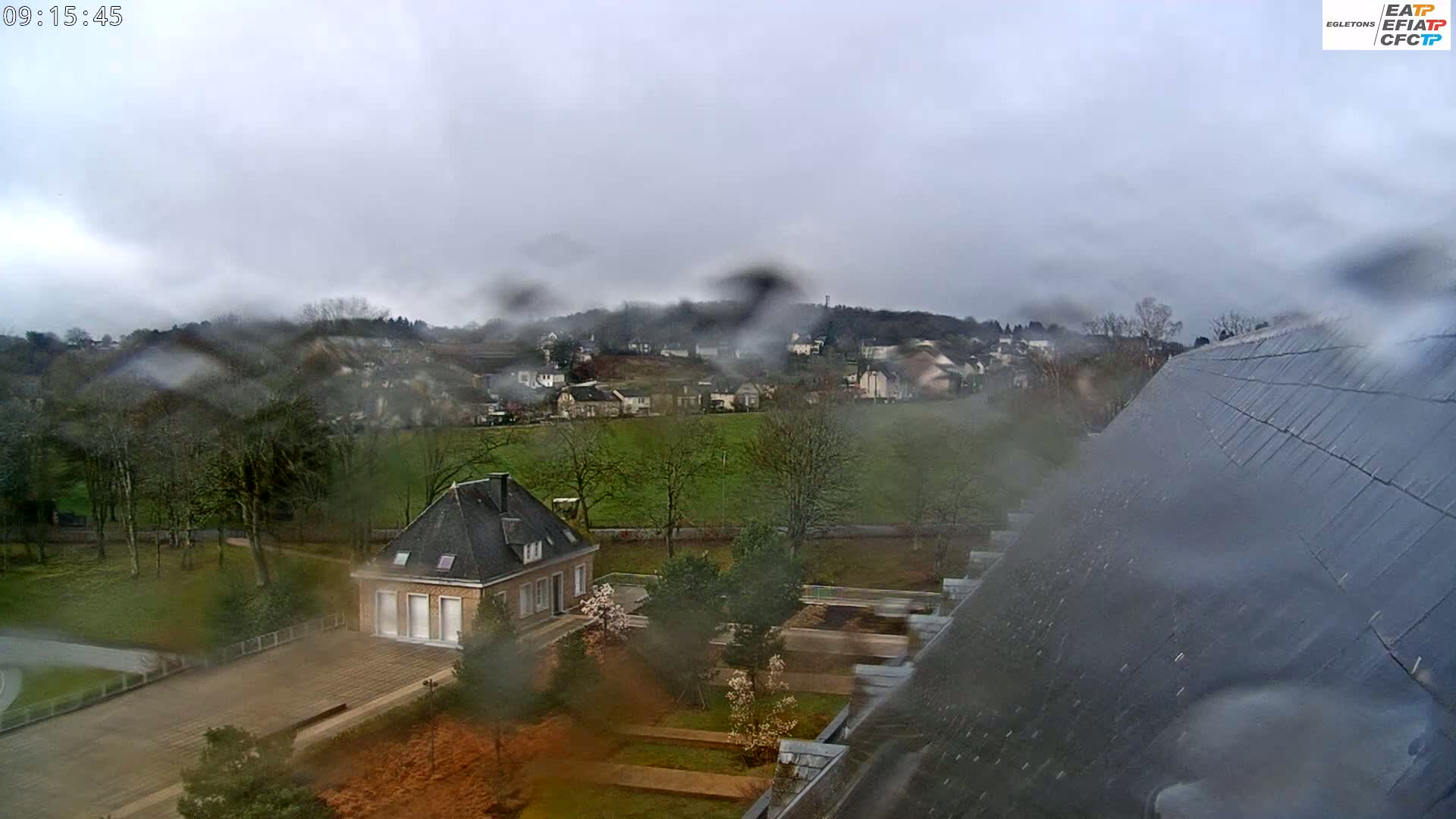 A rain-streaked view shows a small house and a grassy field, beyond which lies a town nestled in a valley under a cloudy sky.