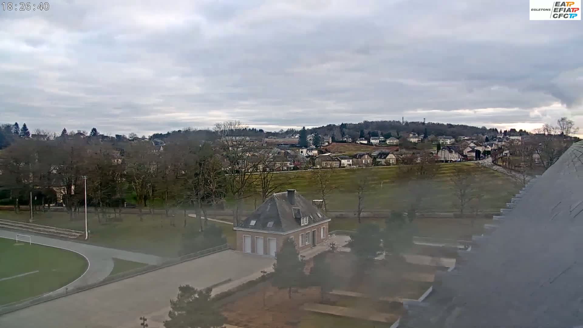 A high-angle, long shot shows a residential area on an overcast day, with a small building in the foreground and a running track visible to the lower left.