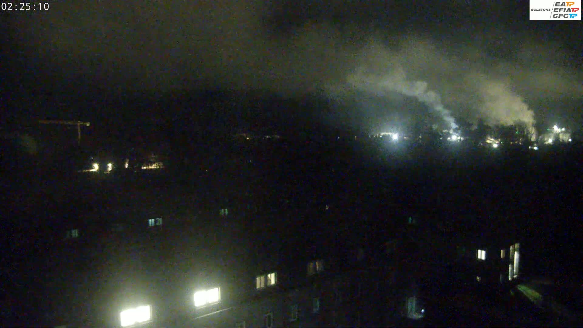 A nighttime, high-angle view shows a dark landscape with faint smoke rising in the distance, illuminated by scattered lights from buildings and possibly industrial areas.