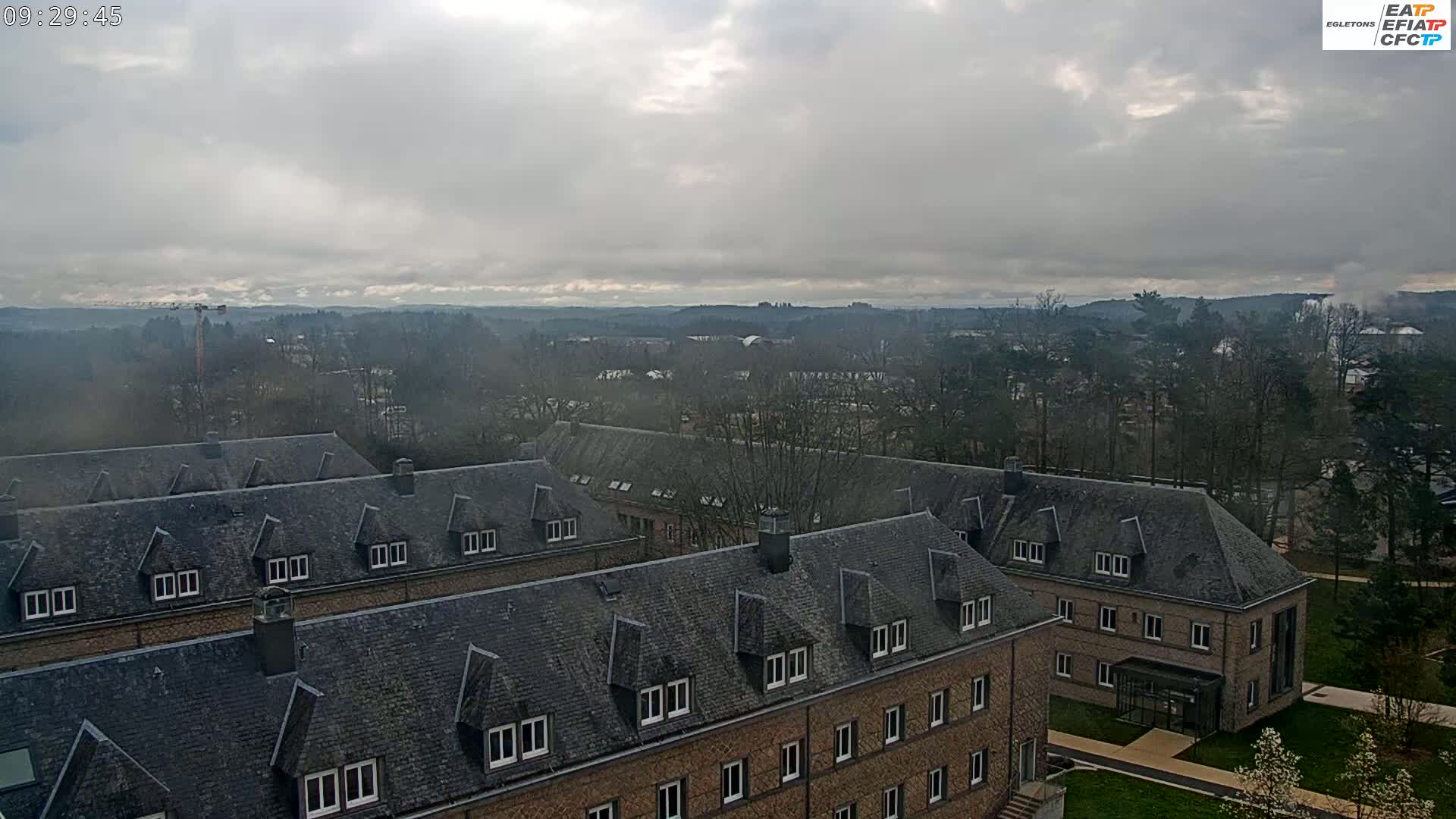 A sprawling complex of low-rise buildings with dark gray slate roofs is situated on a hill overlooking a tree-covered valley under a cloudy sky.