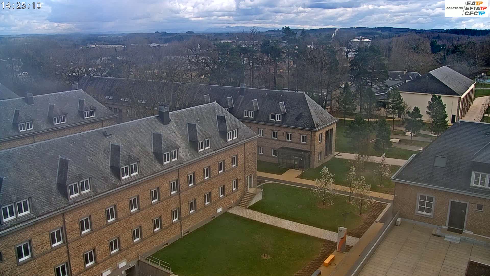 An aerial view of a campus with several stone buildings, grassy areas, and trees under a partly cloudy sky.