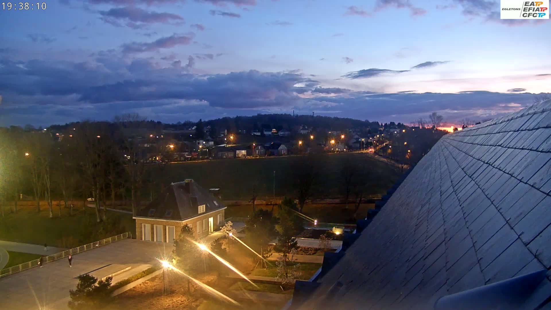 A twilight view shows a town at night with lights illuminated against a partly cloudy sky, overlooking a park-like area with a building and a large sloped roof.