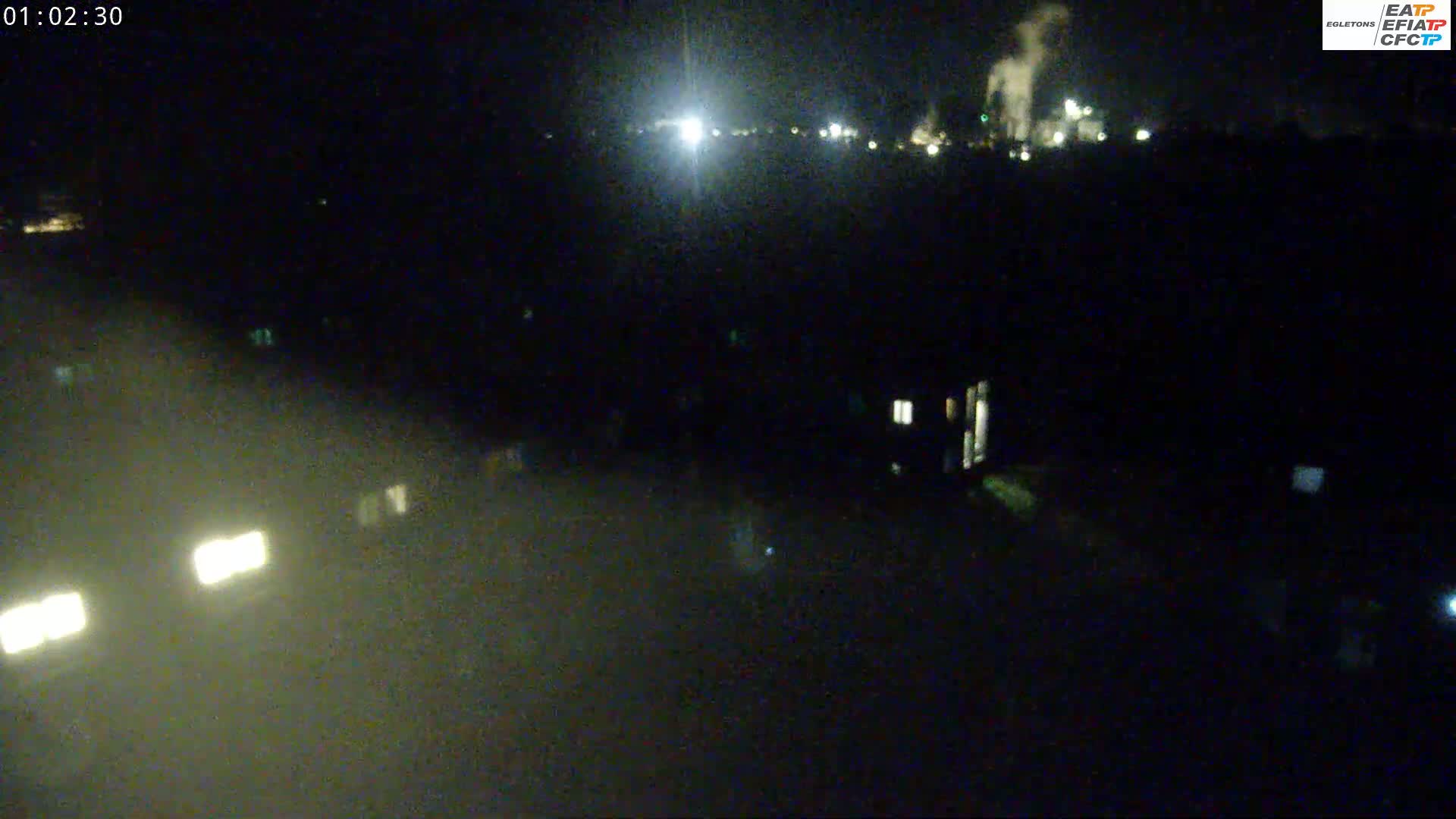 A nighttime, low-light view shows a distant industrial area with lights and smoke, and some nearby illuminated structures and vehicle headlights in the foreground.