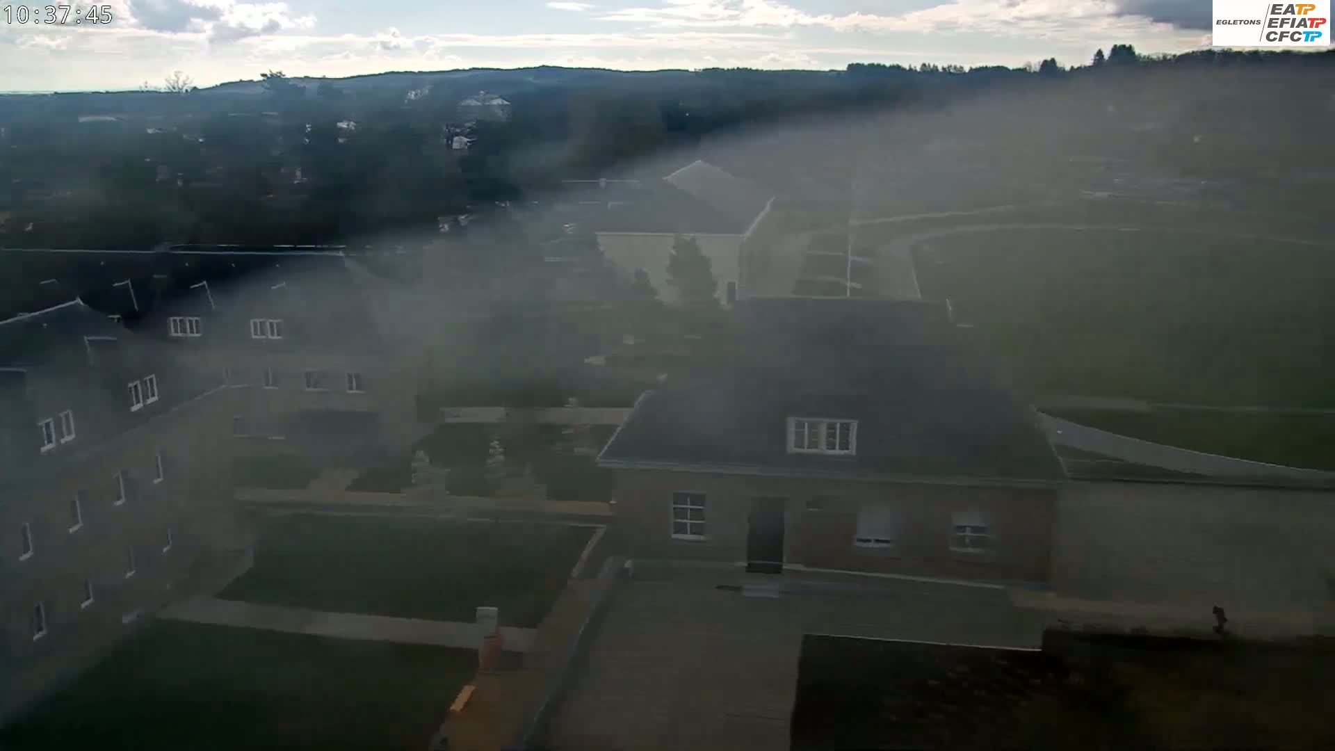 A hazy, partly cloudy day reveals a high-angle view of a residential area with several buildings, including a two-story house and multi-unit dwellings, and green spaces.