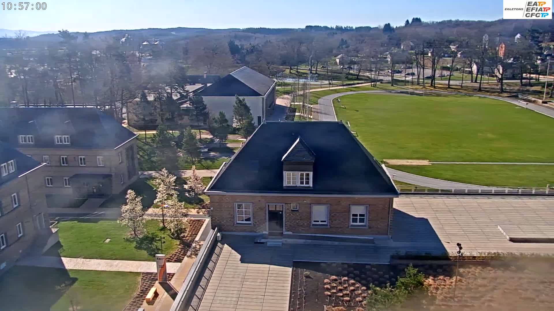 An aerial view of a campus on a sunny day shows several buildings, a grassy field with a running track, and blossoming trees.