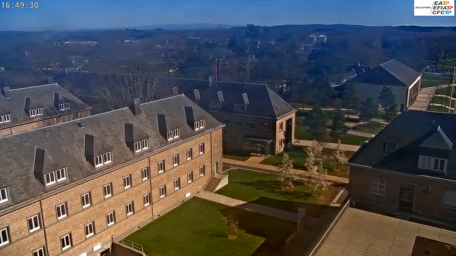An aerial view of several brick buildings on a sunny day, with a sprawling lawn and trees in the foreground, and a distant landscape under a clear blue sky.