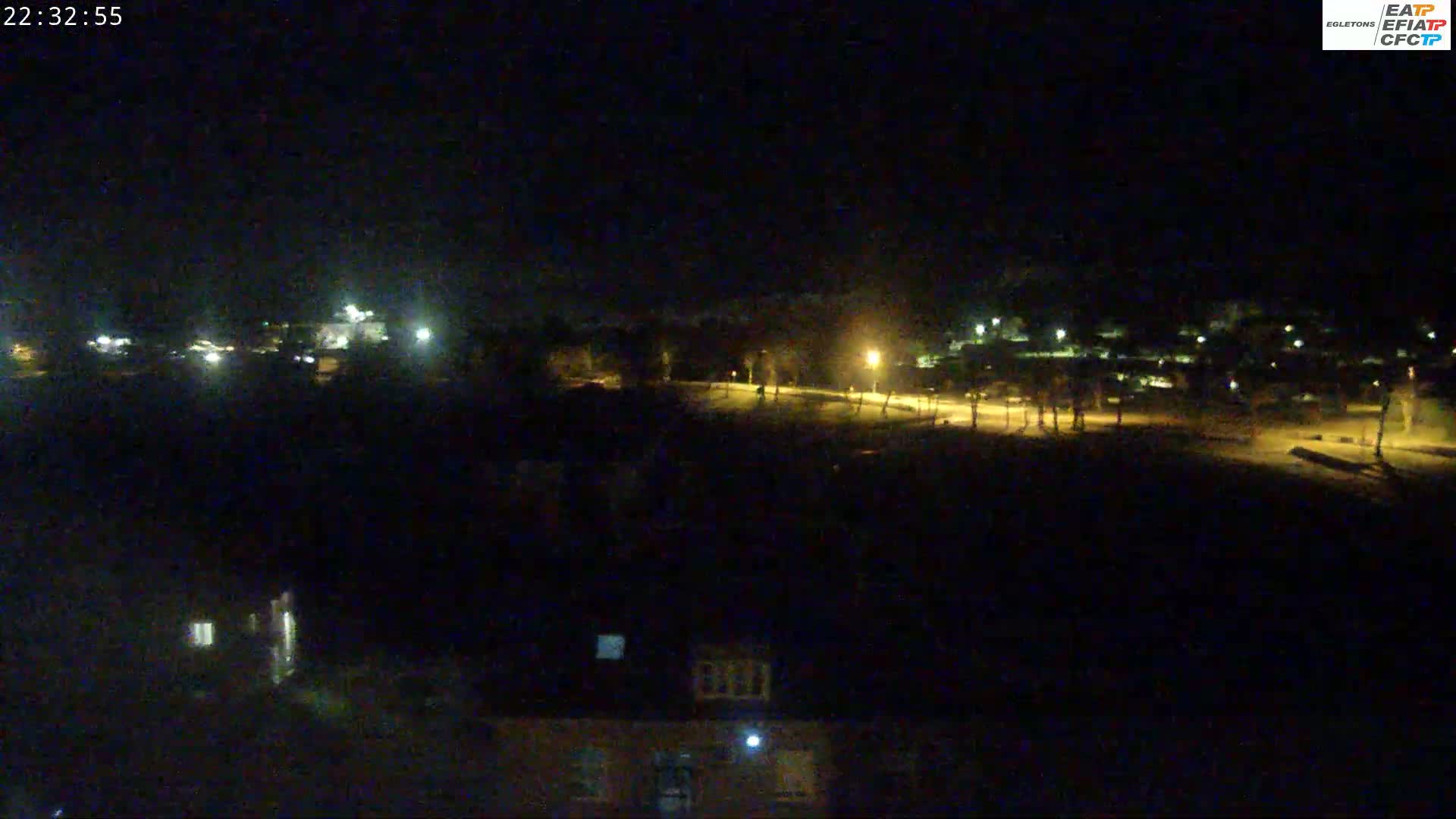 A nighttime long shot shows a dark field illuminated by a few distant lights, with buildings and streetlights visible in the background under a dark sky.