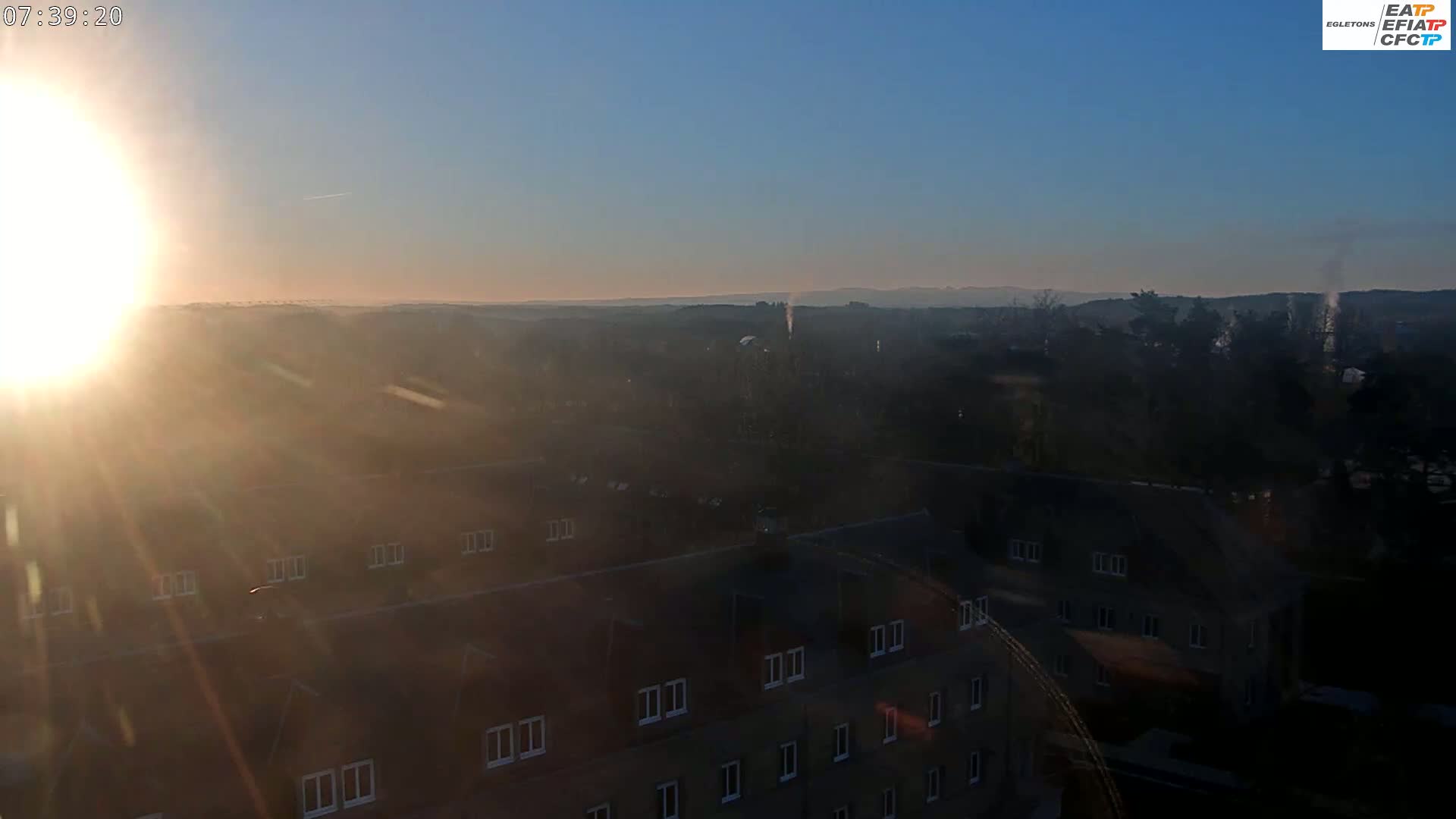 A hazy sunrise illuminates a sprawling landscape of buildings and trees, with a distant horizon visible under a clear, blue sky.