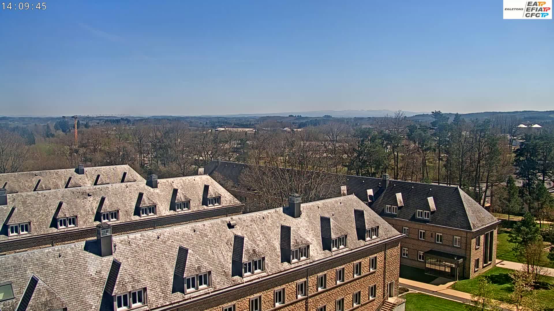 A high-angle view on a clear day shows several buildings with grey slate roofs, surrounded by bare trees and a grassy area, with distant hills visible in the background.