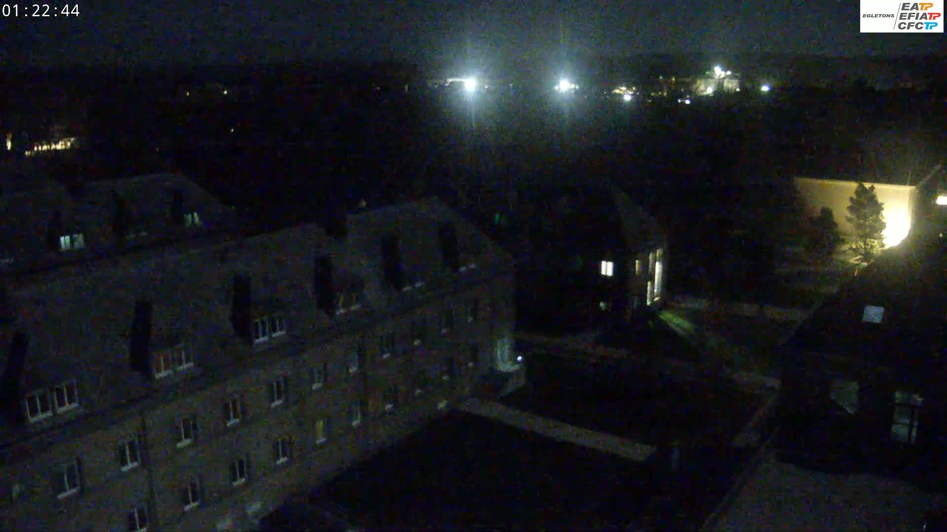 A nighttime aerial view shows several dark buildings with illuminated windows, a courtyard, and distant lights.