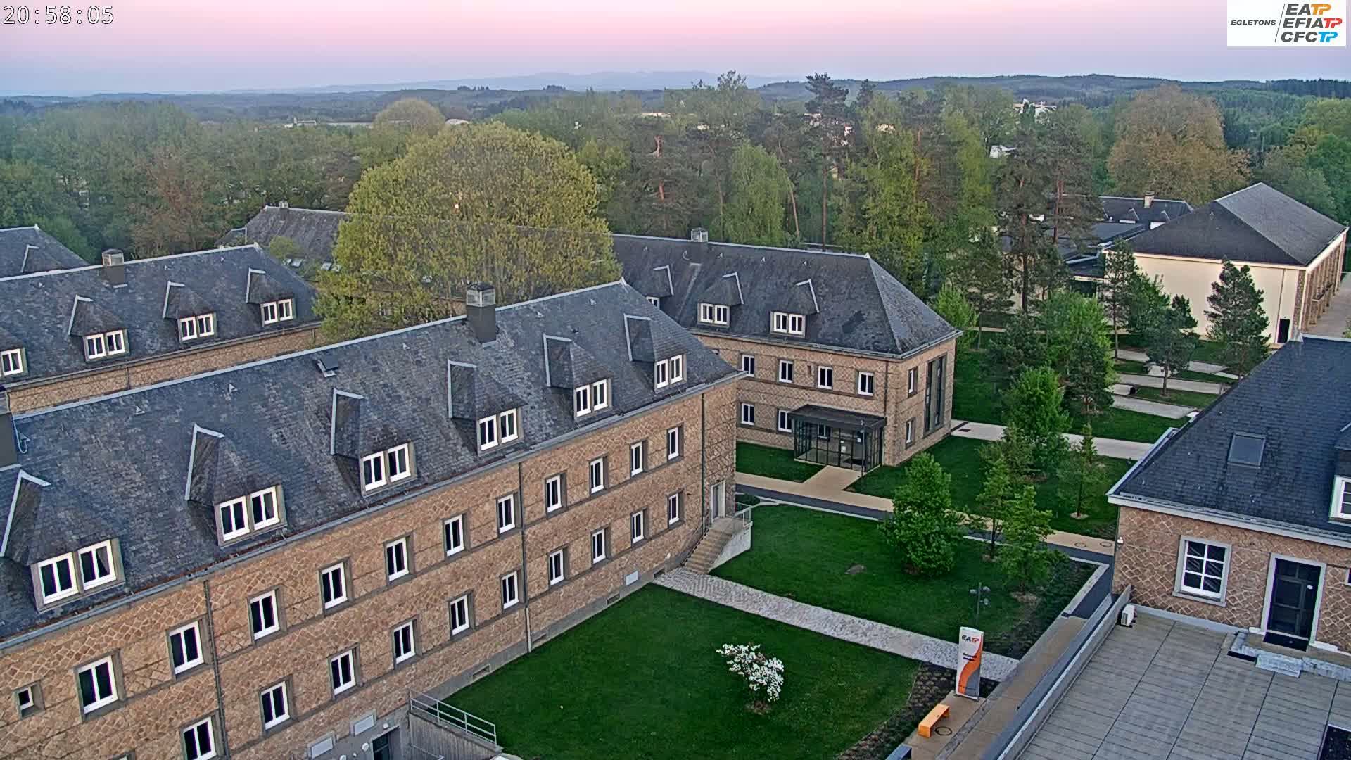 An aerial view shows several low-rise brick buildings with slate roofs on a grassy campus at twilight.