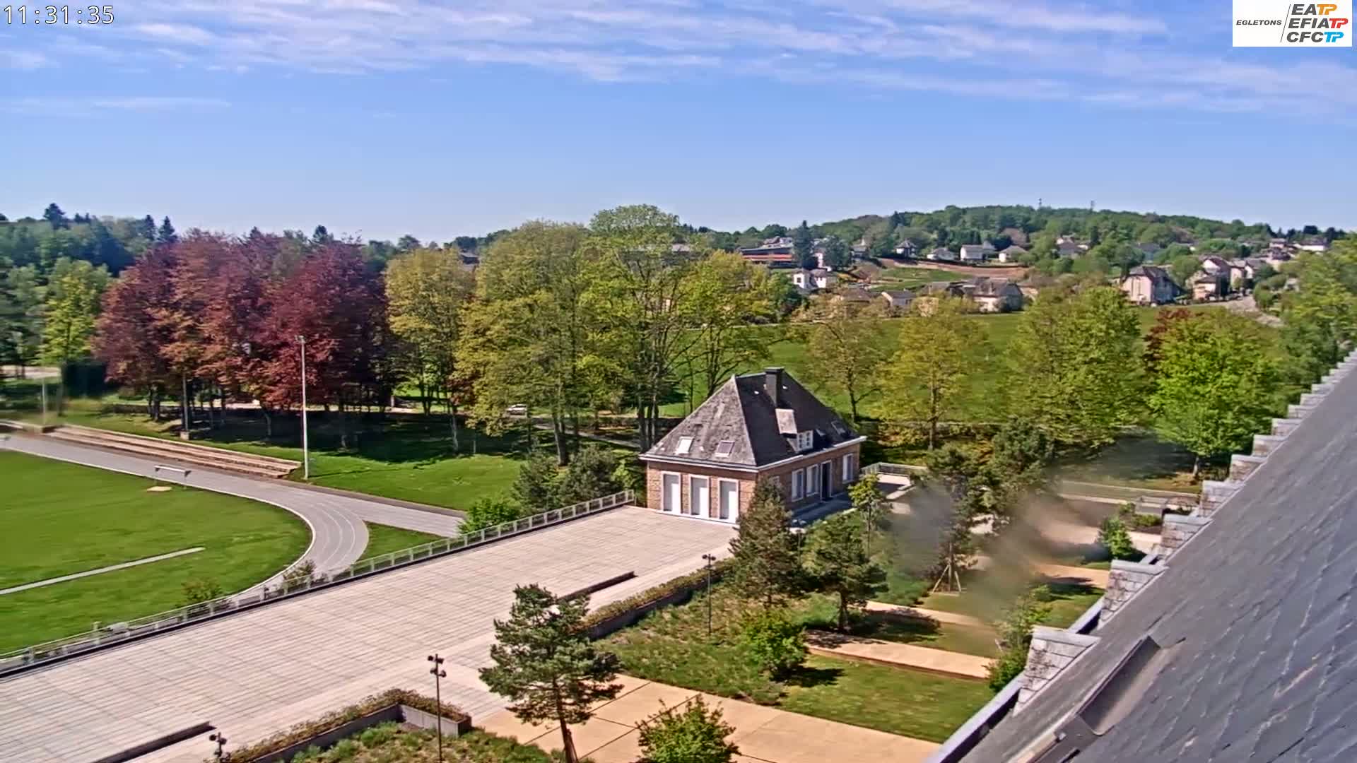 A high-angle view on a sunny day shows a brick building situated in a park-like setting with a running track, lush green lawns, and a small village in the background.