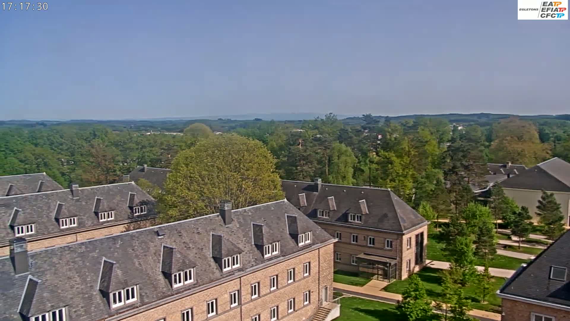 A high-angle view on a clear, sunny day shows several brick buildings with gray roofs surrounded by green trees and manicured lawns.