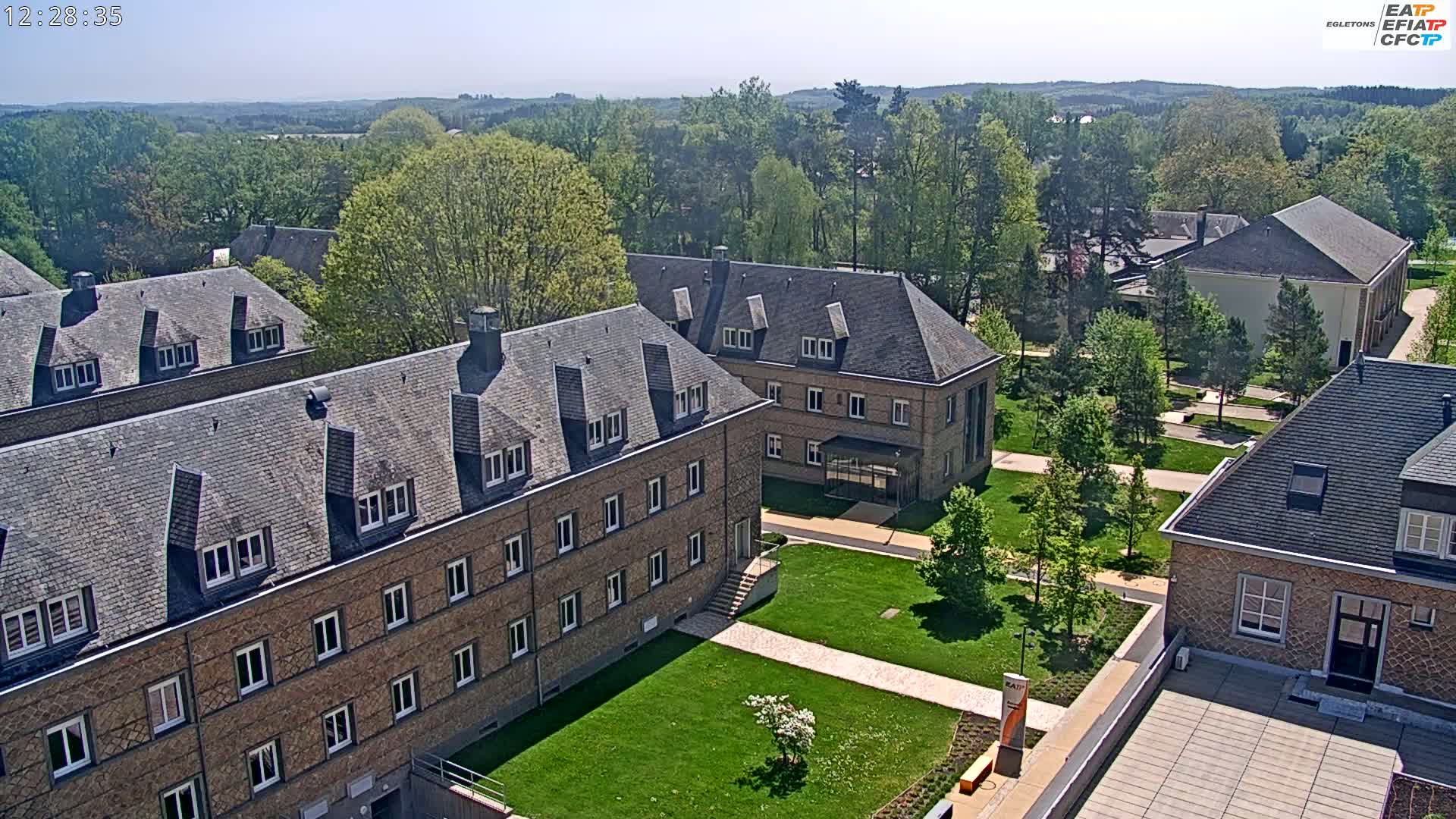 A sunny aerial view of a campus with several brick buildings surrounded by green lawns and trees.