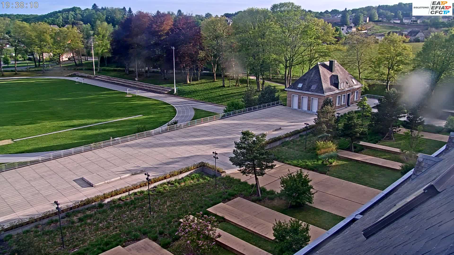 A high-angle view shows a paved area with a running track and grassy field, a small building, and landscaped gardens, all under a mostly clear sky.