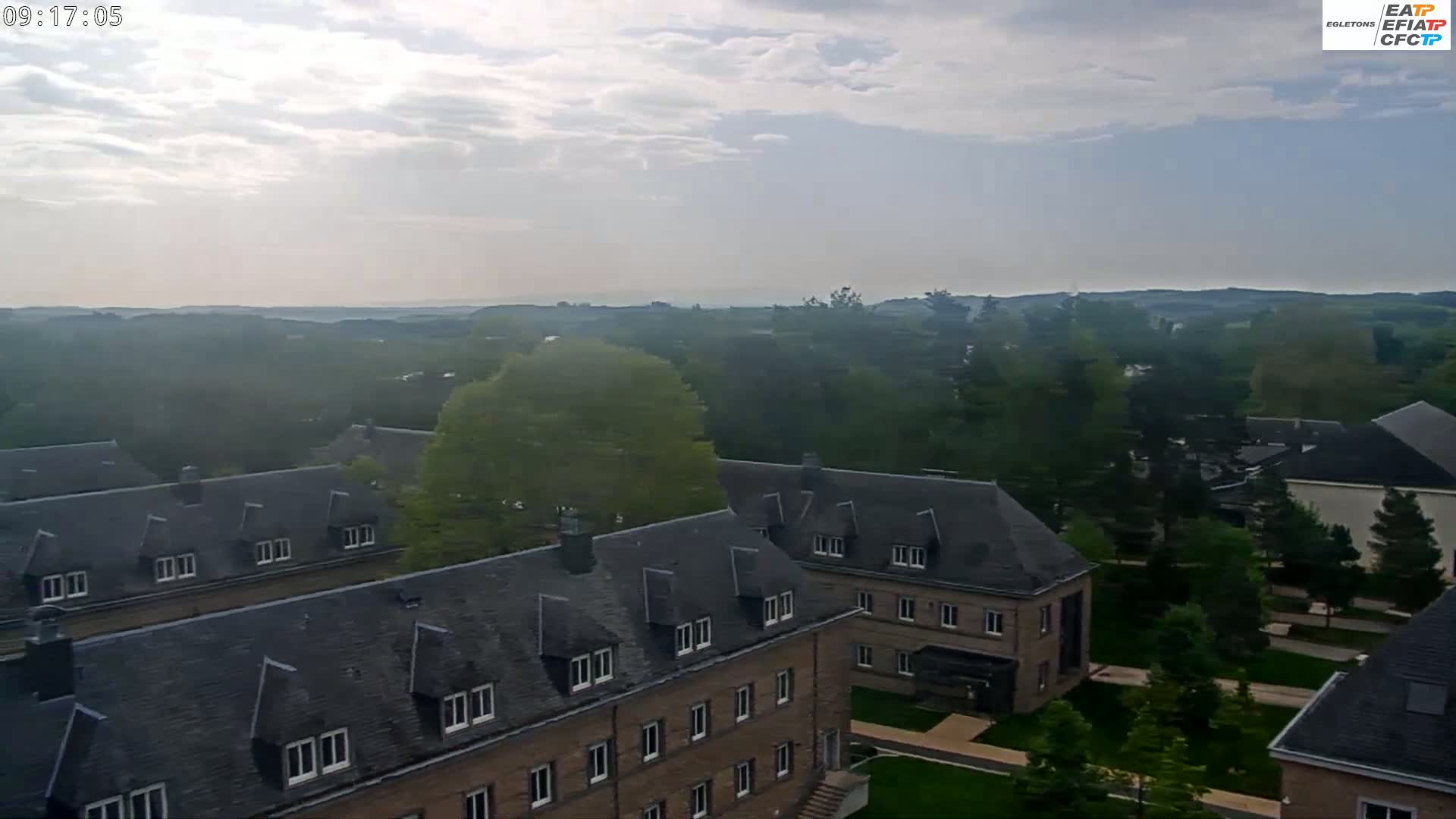 A mostly cloudy sky overlooks a collection of low-slung brick buildings with dark gray roofs, surrounded by green trees and landscaping.