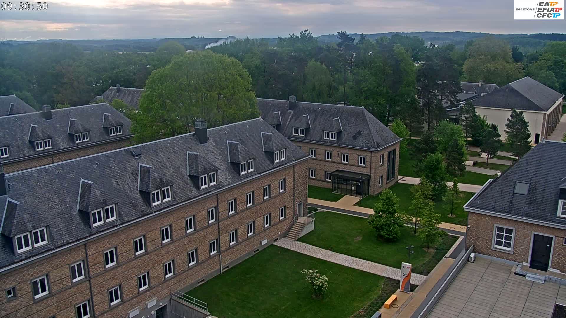 An aerial view shows a campus of low, stone buildings with gray slate roofs under a cloudy sky.