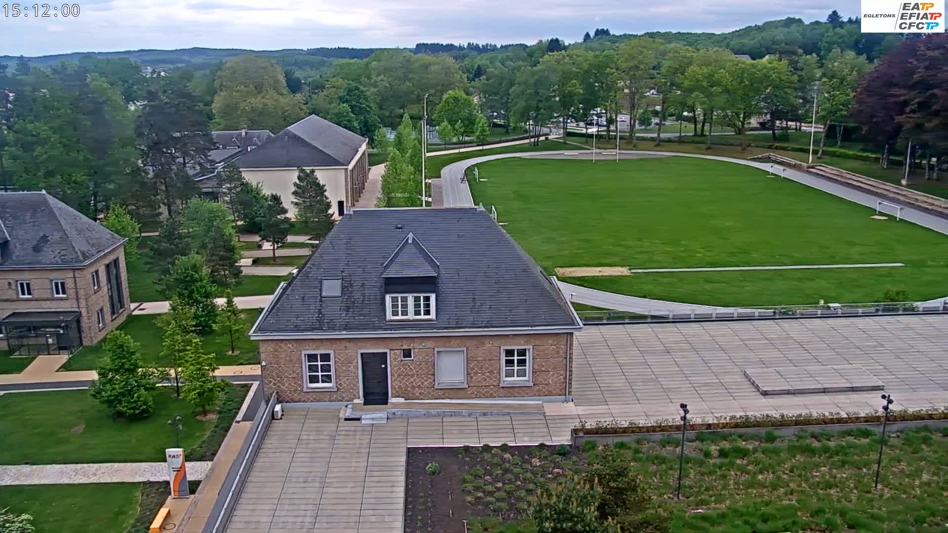An overcast day reveals a campus scene with several stone buildings, a grassy athletic field with a running track, and landscaped areas.