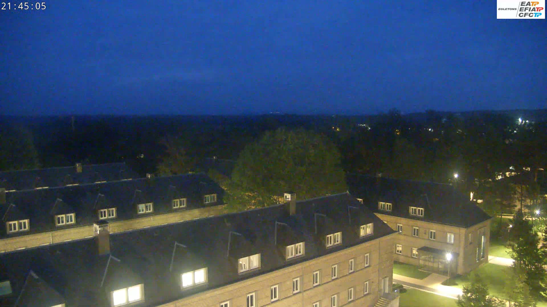 A nighttime, aerial view shows a complex of low-rise buildings with illuminated windows, surrounded by trees and lawns under a dark blue, twilight sky.