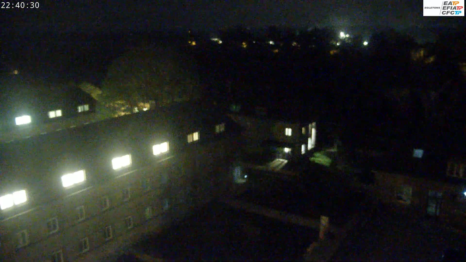 A nighttime, high-angle view shows a cluster of buildings with some illuminated windows, surrounded by dark trees and grounds.