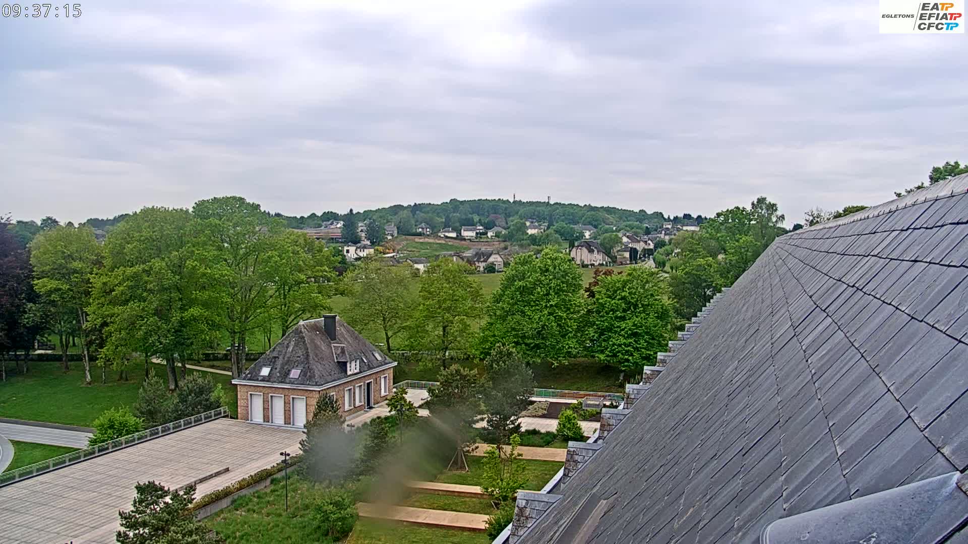 A view of a town under a cloudy sky, showing a small building and a large building's roof in the foreground, surrounded by green trees and lawns.
