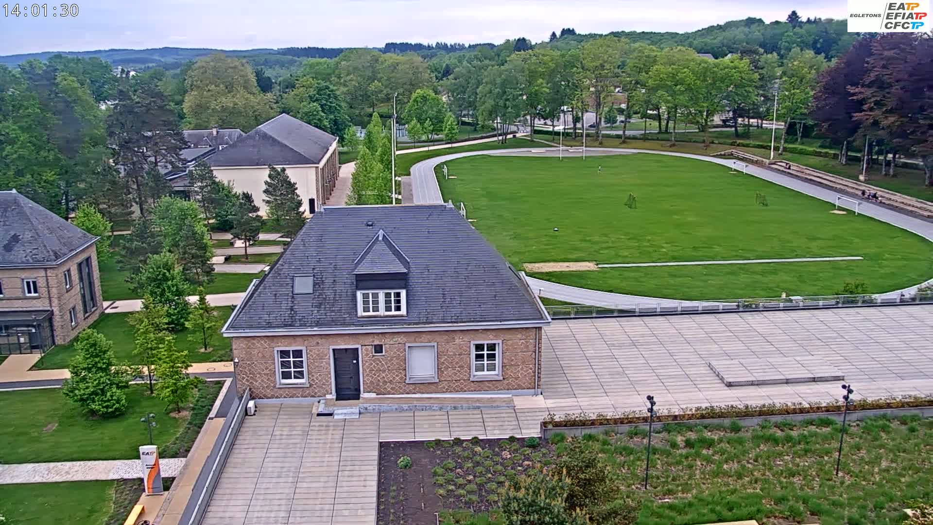 An aerial view shows a large grassy field with a running track surrounding it, several buildings, and lush green landscaping on a partly cloudy day.