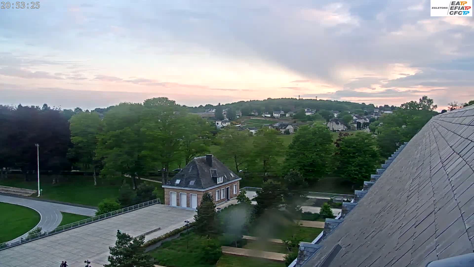 An elevated view shows a small building and a running track in a park-like setting, with a village and hills visible in the distance under a partly cloudy, sunset sky.