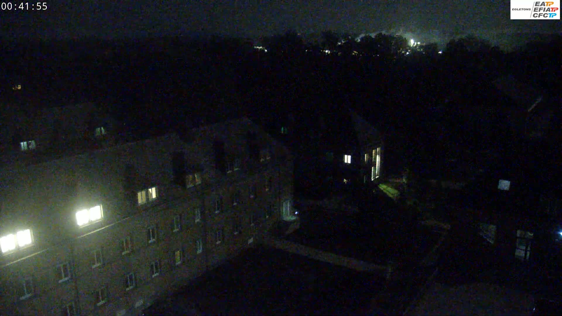 A nighttime, high-angle view shows a multi-story building with some illuminated windows, adjacent to another building and a dark, tree-lined area.