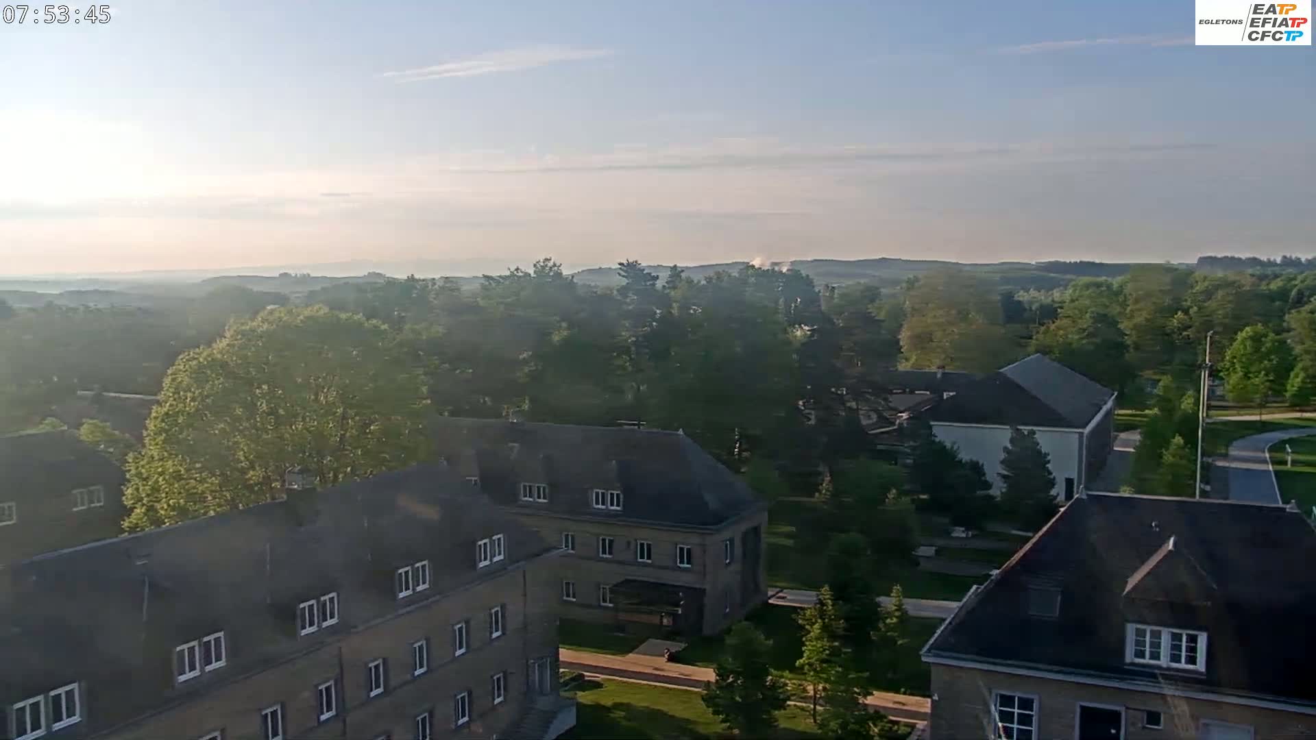 An aerial view shows a hazy morning over a tree-lined landscape with several low-rise buildings.