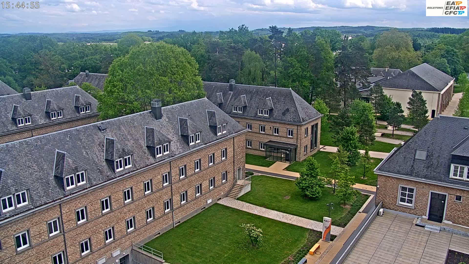 An aerial view of a campus with several stone buildings, green lawns, and trees under a partly cloudy sky.
